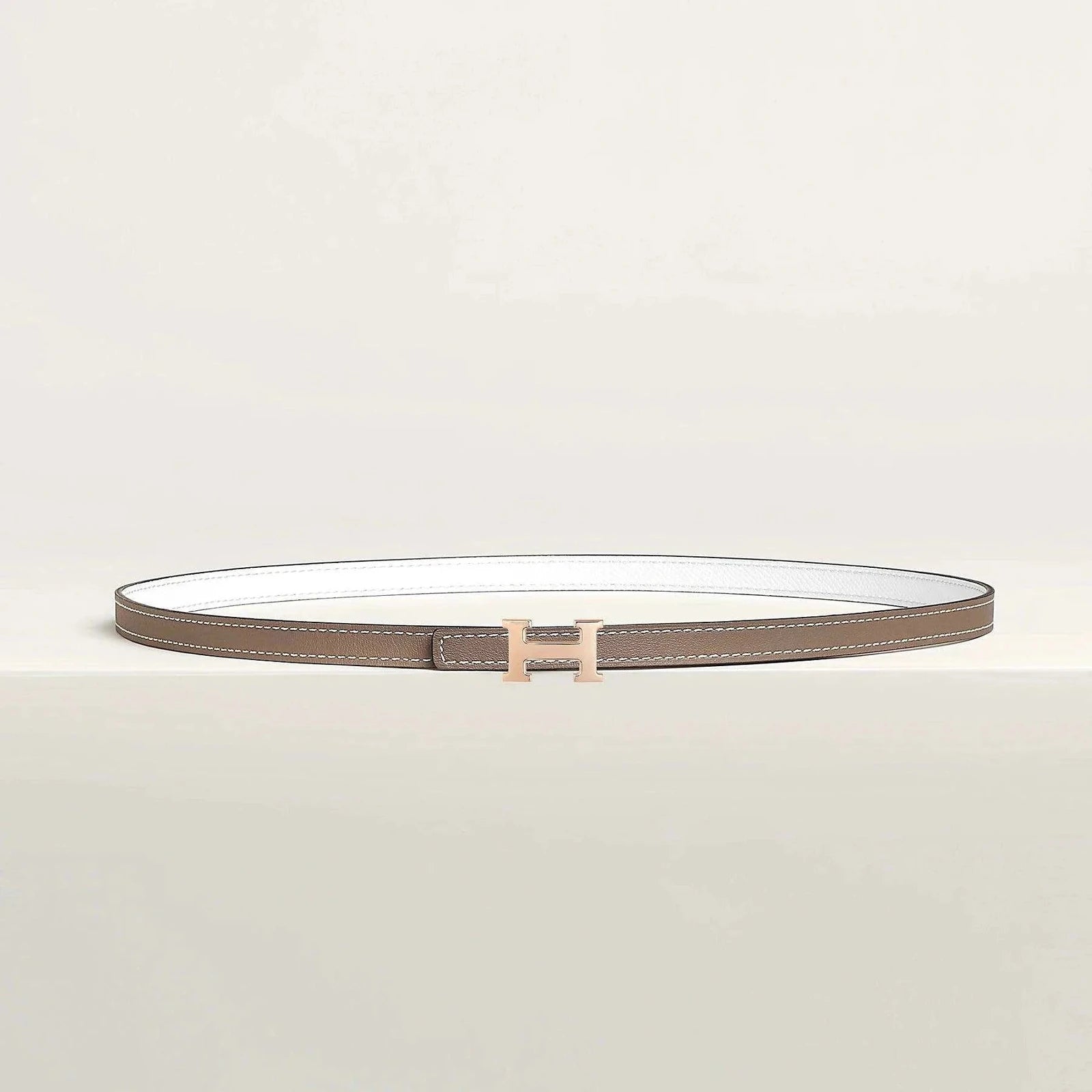 Image of Hermes belt with elegant gold buckle on beige background, showcasing luxury and minimalist design.